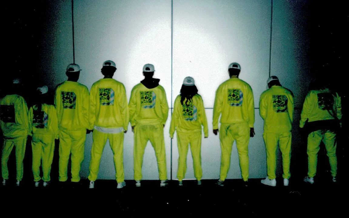 A lineup of Meaningful Existence devotees against a wall. Their backs are turned to the camera, showing off their matching yellow jumpsuits.