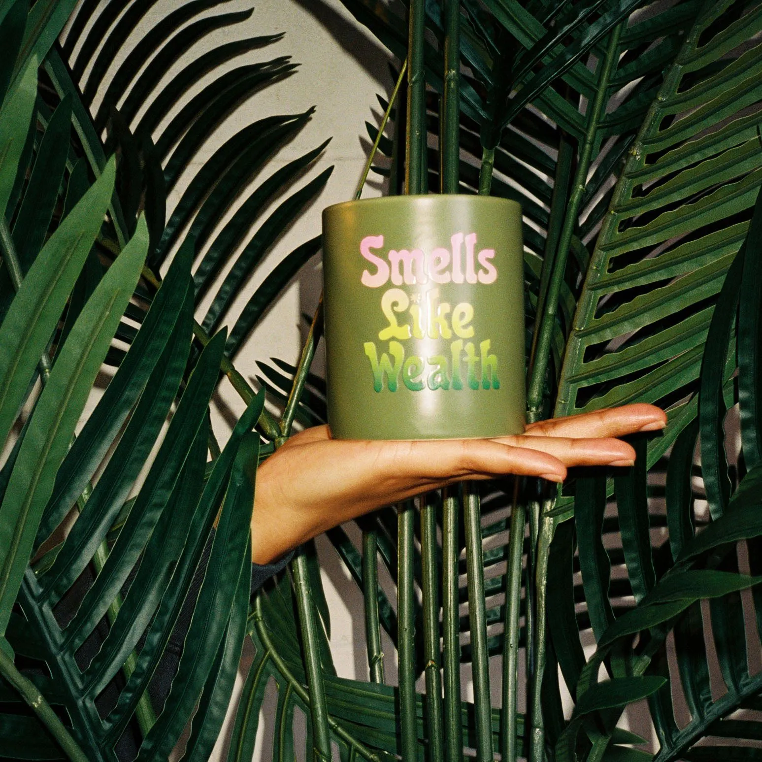 A hand holding a "Smells Like Wealth" candle emerges from some palm fronds.